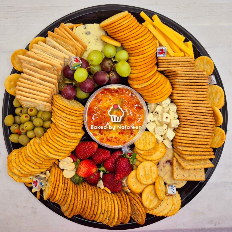 Charcuterie board with crackers, cheese cubes, grapes, olives, strawberries, and a central orange dip labeled 'Baked by Natalie'