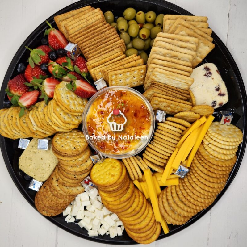 Round black platter loaded with crackers, cheeses, strawberries, grapes, olives, and a central orange dip labeled 'Baked by Nataleen'.