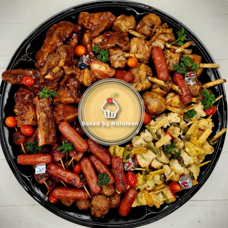 Large black platter of assorted grilled meats, sausages, potatoes, and vegetables arranged around a central creamy dipping sauce.