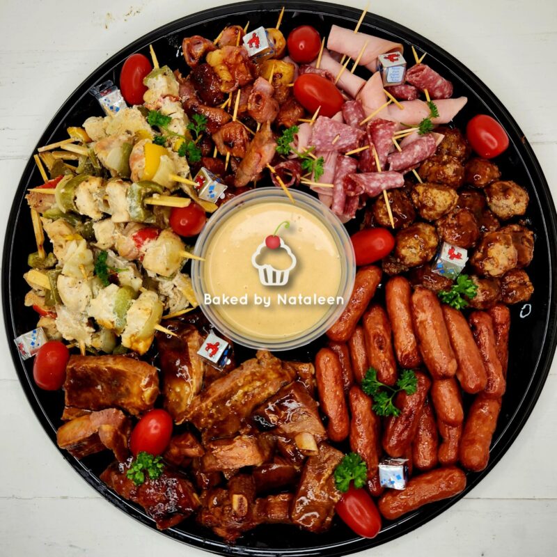 Round black platter with assorted skewered bites arranged around a central dipping sauce cup; includes sausages, meatballs, cheese bites, and cherry tomatoes, with a 'Baked by Nataleen' logo in the center.
