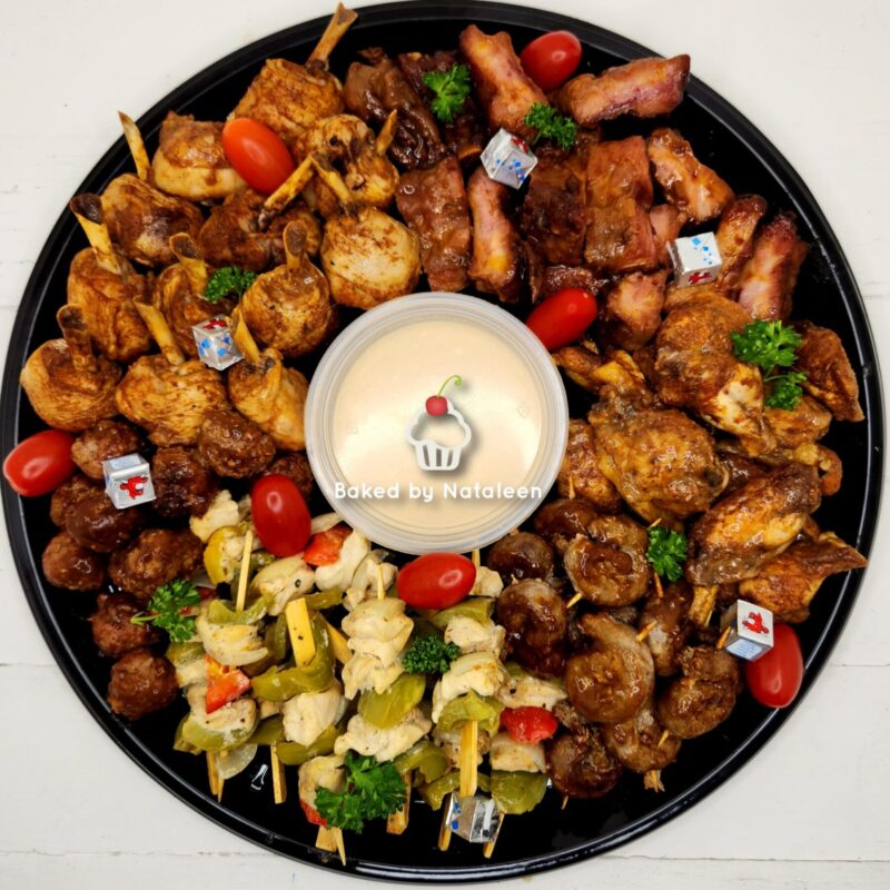 Large round black platter filled with assorted baked appetizers—chicken skewers, meatballs, bacon-wrapped bites, veggie skewers, cherry tomatoes, and a central dipping cup labeled 'Baked by Nataleen'.