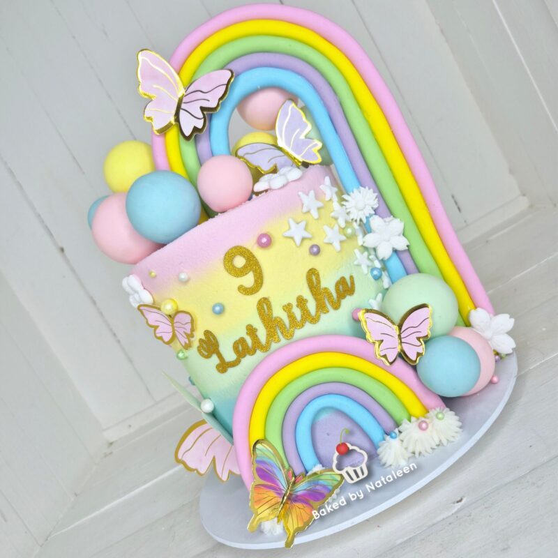 Pastel birthday cake with rainbow arches, pastel spheres, butterflies, and the name 'Lachlan' with the number 9 in gold glitterartifacts—baked by Nataleeon styling included.