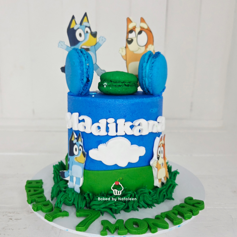Two-tier birthday cake with blue and green fondant, cartoon dog figures, green macarons on top, cloud cutouts, and the name 'Madikane' on the side.