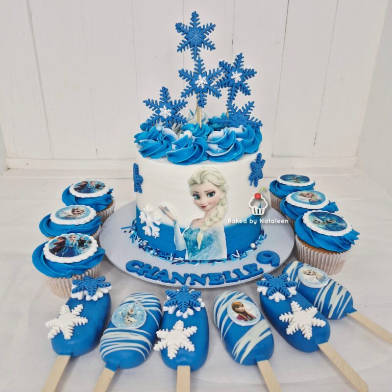 Elsa-themed birthday cake with blue swirls, snowflakes, and a fondant Elsa image, surrounded by blue cupcakes and snowflake popsicles on a white table.