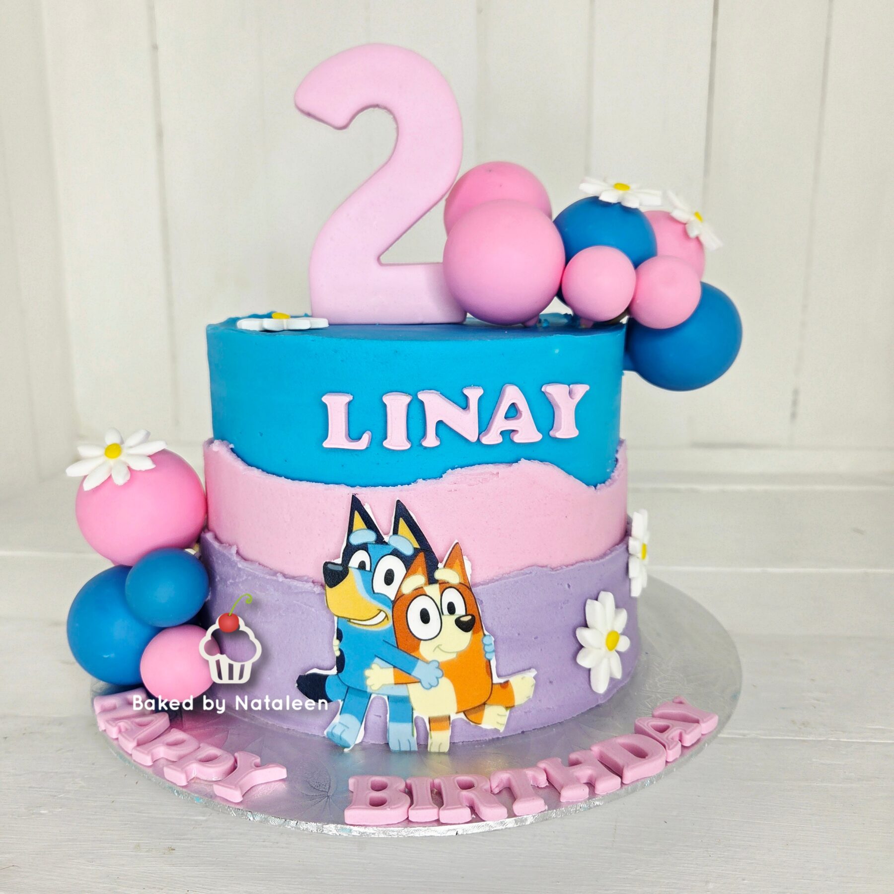 Two-tier pastel birthday cake with a large pink '2' topper, blue and pink layers, balloons, flowers, and cartoon characters on the side for a child’s celebration.