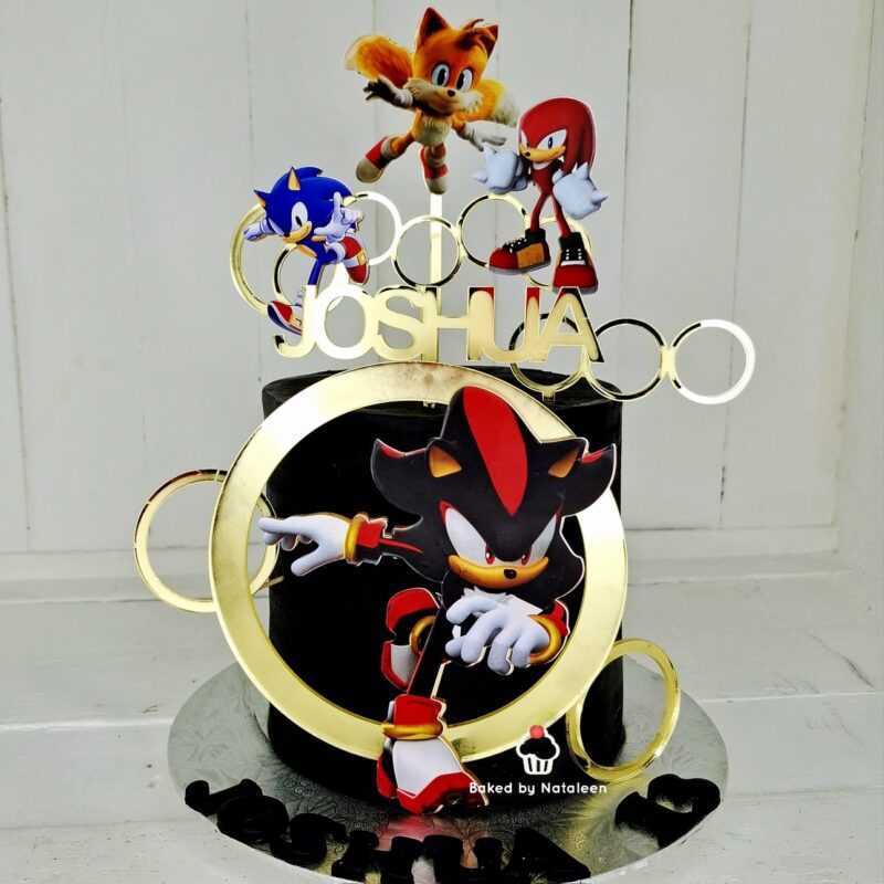 A birthday cake with Sonic the Hedgehog decorations, gold rings, and the name Joshua on top, set on a white background.