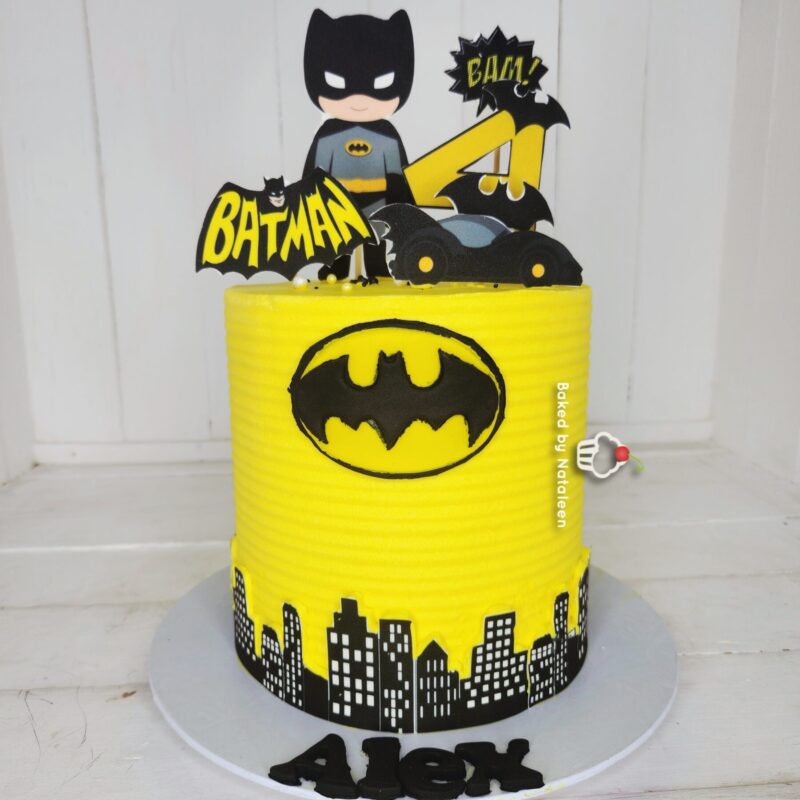 Batman-themed birthday cake with yellow fondant, Batman logo, city skyline border, and dark superhero toppers; name 'Alex' on base.