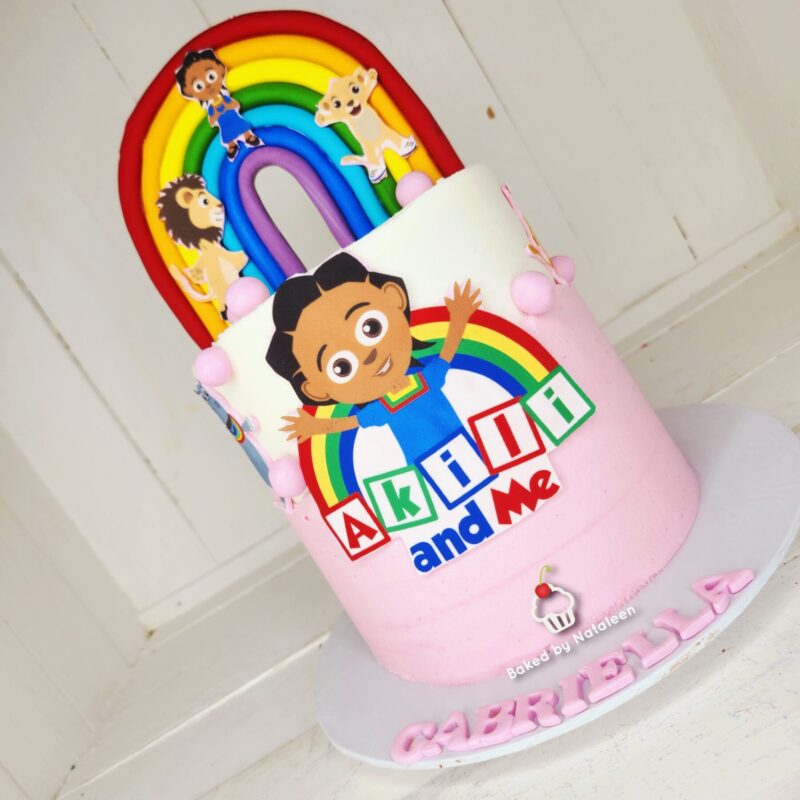 Pink birthday cake with a large rainbow and cartoon child on the front; the cake shows 'AKODI and Me' letters and 'GABRIELLA' on the base, baked by Nataleen.