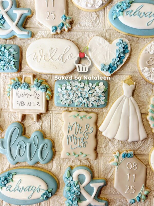 Assorted wedding-themed cookies in blue, white, and beige with messages like 'love', 'we do', and 'happily ever after' along with a small dress-shaped cookie, arranged on a tray.