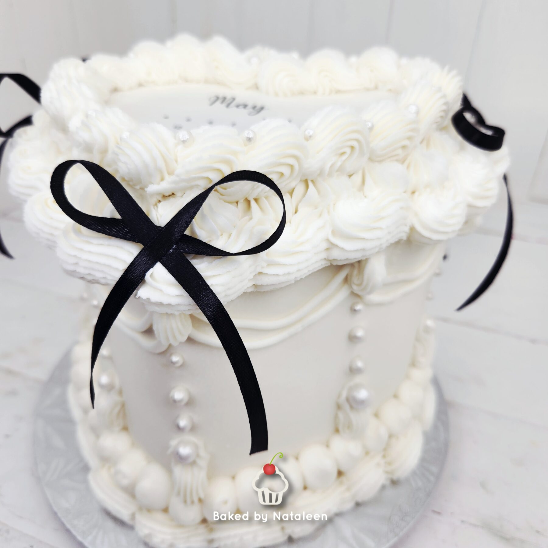 White cake decorated with piped rosettes, pearl beads, and a black satin ribbon on a white background, showing a watermark at the bottom reading 'Baked by Nataleen'.