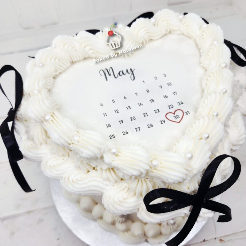 White frosted cake with piped borders and a May calendar showing the 30 circled in red, ribbons at the sides, Baked by Nataliee label visible.