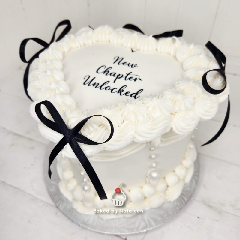 White frosted cake with piped cream border, black ribbons, and a plaque reading 'New Chapter Unlocked' on top