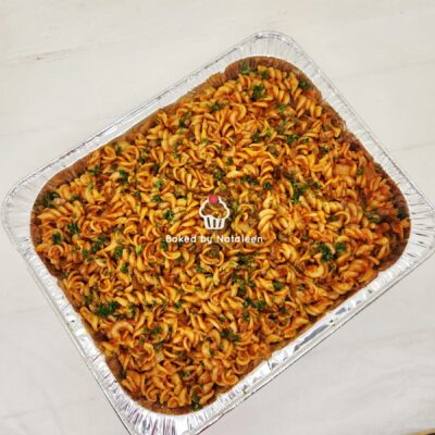 Tray of baked rotini pasta with tomato sauce and chopped parsley, in a disposable aluminum tray.