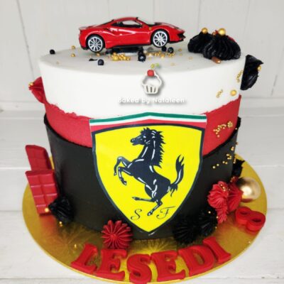 Two-tier Ferrari-themed cake: black and red fondant with a large yellow Ferrari shield on the side, white top with a red toy sports car and gold sprinkles, red and black decorations, and red fondant letters around the base spelling LES EDOL.