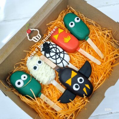 Box of Halloween cake pops on orange shredded paper: green monsters, a white mummy, a black bat, a spiderweb, and a pumpkin-shaped pop in a gift box.