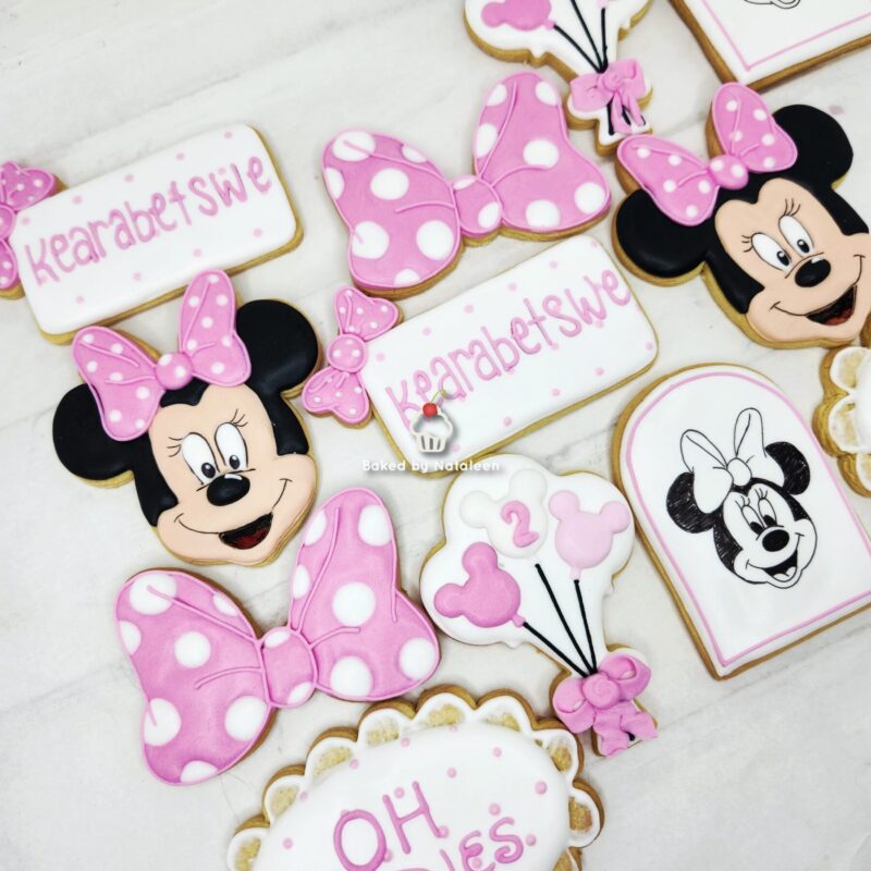 Cookies decorated with Minnie Mouse faces, pink polka-dot bows, and pink lettering on white icing, pastel cupcake theme (baked items shown)