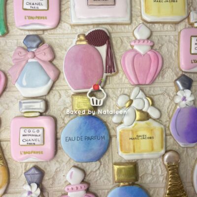 Assorted pastel perfume-bottle cookies with bows and flowers on a light brick backdrop, watermark 'Baked by Nataleen'