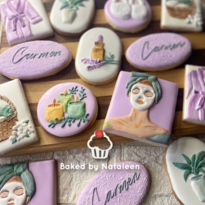 Assorted pastel cookies with spa and tropical designs on a wooden board, including face mask figure and candles.