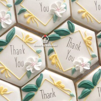 Hexagonal cookies iced white with yellow borders and green leaves, each saying 'Thank You' in slim text amid white flowers; a watermark reads 'Baked by Nataleen' across the tray.