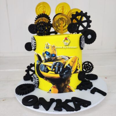 Yellow cake with Transformer character image, metallic gears around and 'Baked by Nataleen' — a birthday cake centerpiece with black sugar gears.