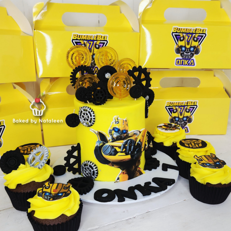Yellow Bumblebee Transformers birthday cake with gear toppers and a large Bumblebee image, surrounded by matching cupcakes and yellow gift boxes in the background.