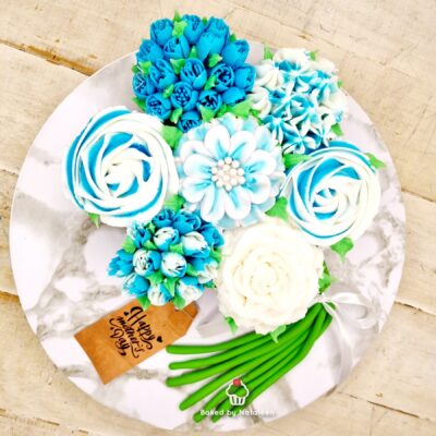 Buttercream floral bouquet on a marble cake board in blue, white, and green with a 'Happy Birthday' tag and green stems showing at the bottom