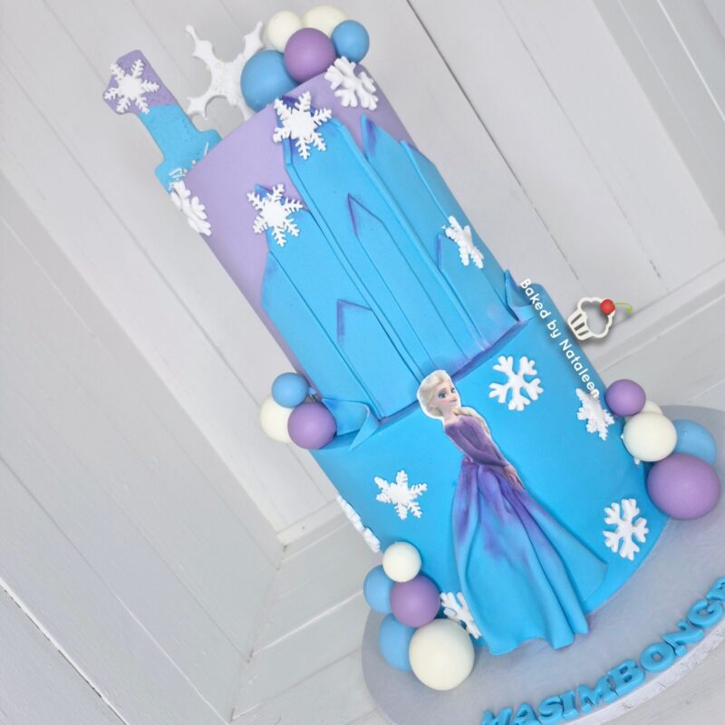 Two-tier Frozen-themed birthday cake in blue and purple with snowflakes, a castle silhouette on top, and an Elsa figurine with decorative purple and white spheres around the base.