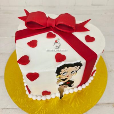 White fondant square cake with a large red bow and diagonal red ribbon, red heart decorations, Betty Boop cutout on the side, white pearl border on a gold cake board