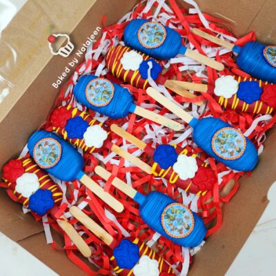 Colorful candy-pop party favors in a box: blue wrappers, yellow stripes, wooden sticks, red/white confetti, with a bakery label visible.