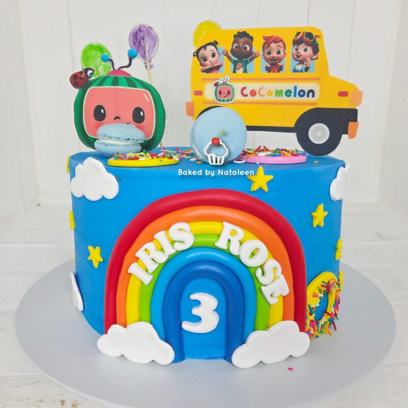 Colorful birthday cake with a rainbow arch, clouds, and a yellow bus topper featuring cartoon kids; reads 'Trix Rose' and shows the number 3 (age).