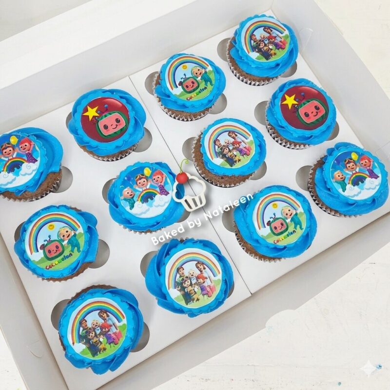 Twelve cupcakes with bright blue frosting in a white box, each topped with round edible images of a rainbow and cartoon children.