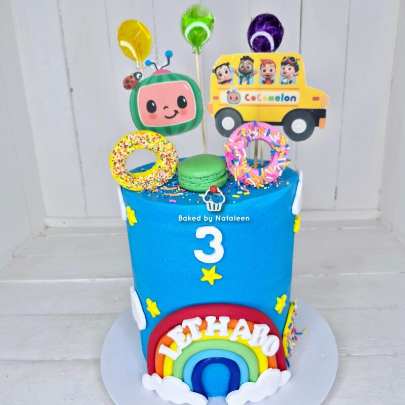 Two-tier birthday cake with blue top, rainbow fondant bottom, the number 3, rainbow arch and clouds, donuts and macaron decorations, CoComelon bus toppers; 'Baked by Nataleen' text.
