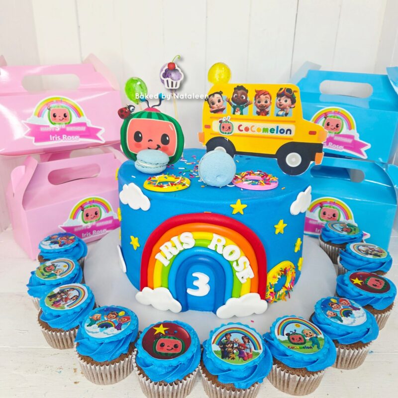 Colorful birthday cake with a rainbow door, clouds, and a blue base, surrounded by blue cupcakes; 'IRIS ROSE' 3rd birthday theme with CoComelon bus topper.