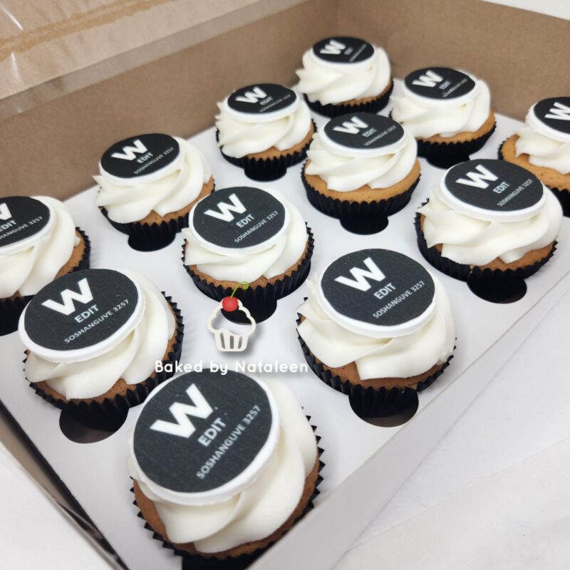 Box of vanilla cupcakes with white swirled frosting and black round toppers labeled 'W EDIT' arranged in a white bakery box.
