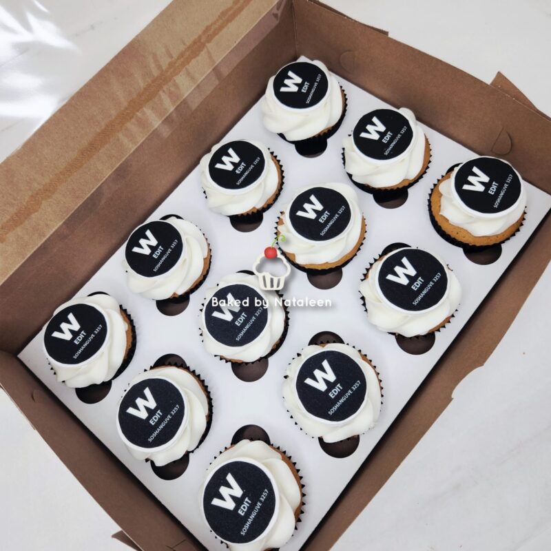 Twelve cupcakes with white frosting and black W EDIT toppers arranged in a white bakery box.