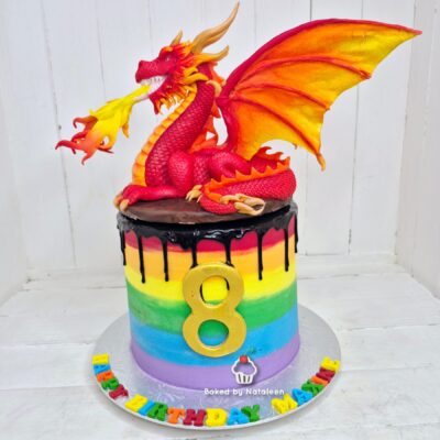Rainbow birthday cake with black drip, a gold '8', and a red dragon figurine perched on top.