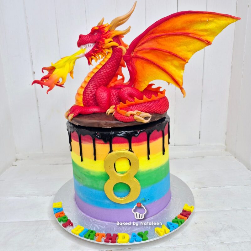 Rainbow birthday cake with black drip, a gold '8', and a red dragon figurine perched on top.