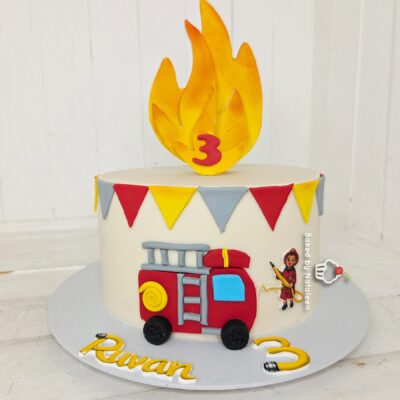 Birthday cake with a large yellow flame topper, a red fire truck decoration, and colorful bunting for a 3rd birthday on a white cake board.