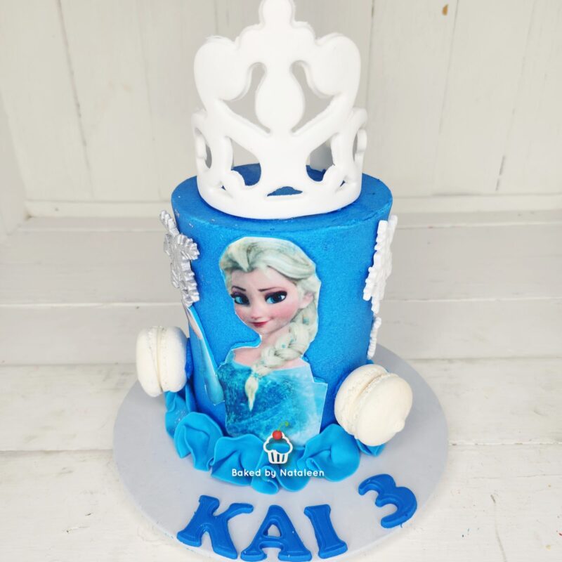 Blue birthday cake featuring Elsa from Frozen with a white crown topper and white macarons on the sides, age 3 and name Kai 3 on the base.