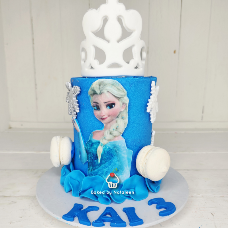 Elsa-themed birthday cake with blue fondant, a white decorative topper, white macarons on the sides, snowflake accents, and blue ruffles at the base (base letters read 'KAL3').