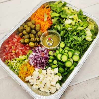 Colorful chopped vegetable tray with olives, feta, cucumbers, tomatoes, onions, peppers, shredded carrots, greens, and a central dip in a foil tray.