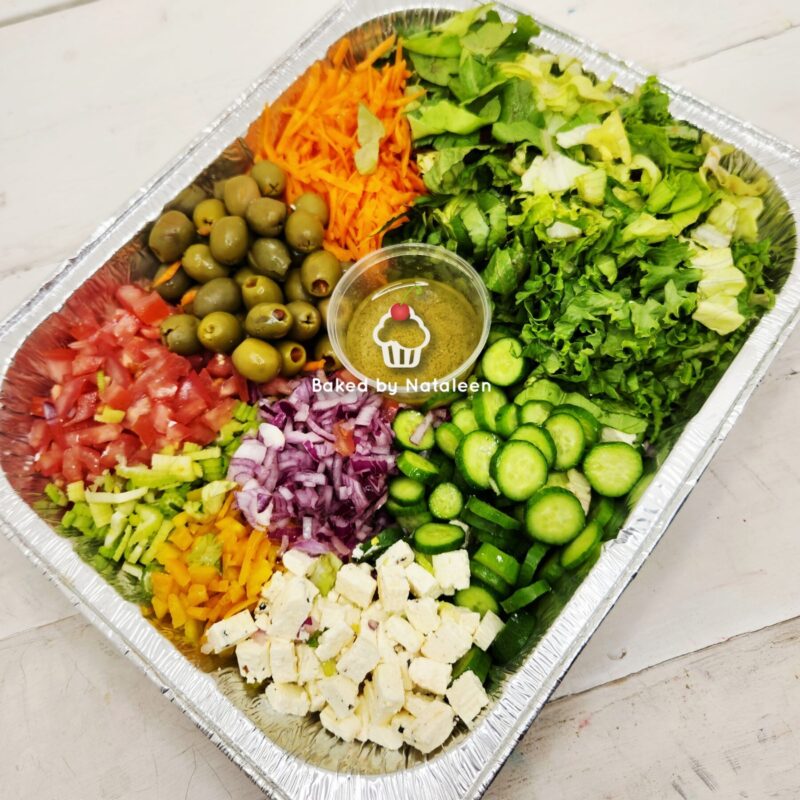 Colorful chopped vegetable tray with olives, feta, cucumbers, tomatoes, onions, peppers, shredded carrots, greens, and a central dip in a foil tray.