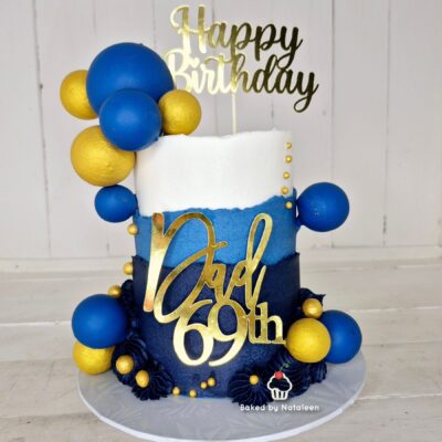 Two-tier birthday cake in white, blue, and navy with a gold 'Dad 69th' topper, 'Happy Birthday' sign, and blue and gold balloons.