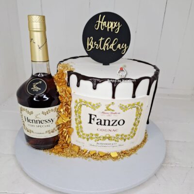 Birthday cake with white frosting and chocolate drizzle sits beside a bottle of Hennessy Very Special cognac on a white cake board, for a birthday celebration.