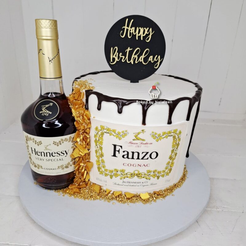 Birthday cake with white frosting and chocolate drizzle sits beside a bottle of Hennessy Very Special cognac on a white cake board, for a birthday celebration.
