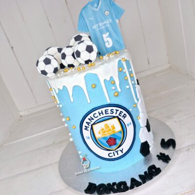 Manchester City themed birthday cake in blue with footballs on top, a small jersey topper, and the club crest on the side.