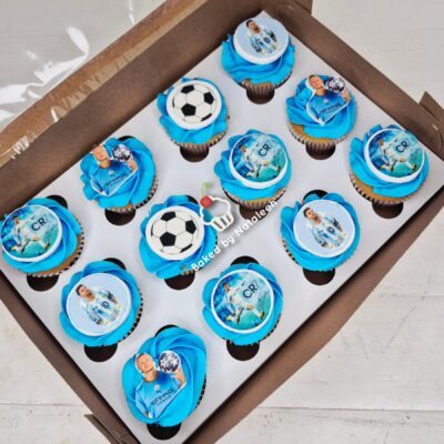 Twelve soccer-themed cupcakes with bright blue icing and edible toppers featuring players, arranged in a brown box with a football motif.