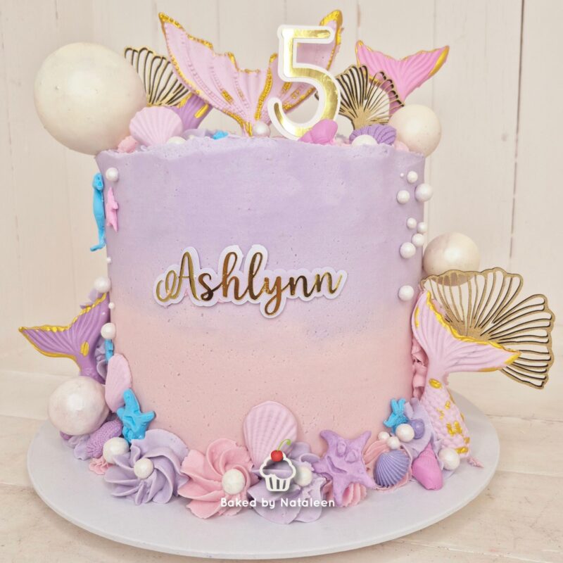 Pastel purple-pink birthday cake with 'Ashlynn' name and a gold 5 topper, sea shell decorations, and mermaid-themed accents.