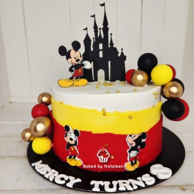 Two-tier birthday cake in white, yellow, and red with a Mickey Mouse topper and castle silhouette; colorful balloons on the side.