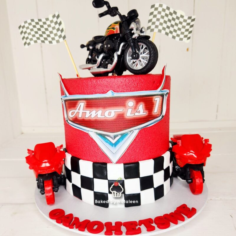 Red racing-themed birthday cake with black-and-white checkerboard band, toy motorcycles on top, and checkered flag accents on the sides and base.